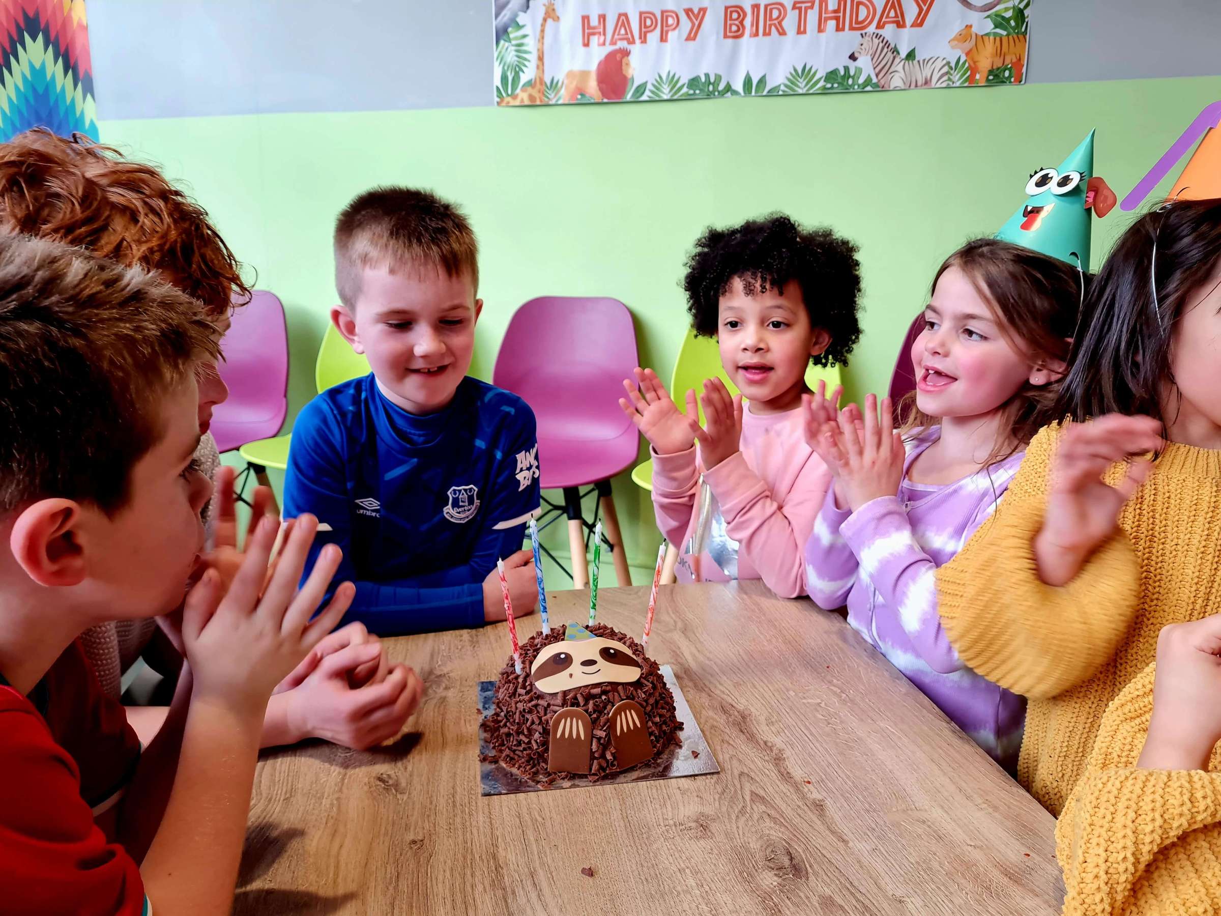 a group of children sitting at a table with a birthday cake, clapping