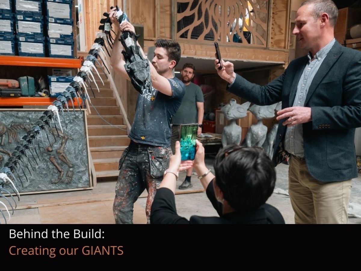 A man and a woman standing in front of a replica skeleton of Titanoboa in production, taking photos on their mobile devices