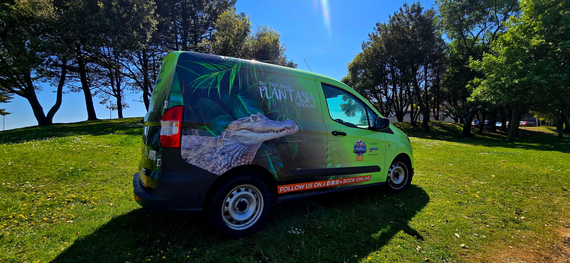 The Plantasia on the Road van with it's new green wrap, parked on the grass with trees and blue sky in the background
