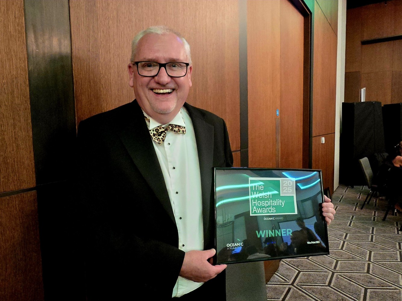 Man in a suit holding an award plaque, smiling in a wood-paneled room.