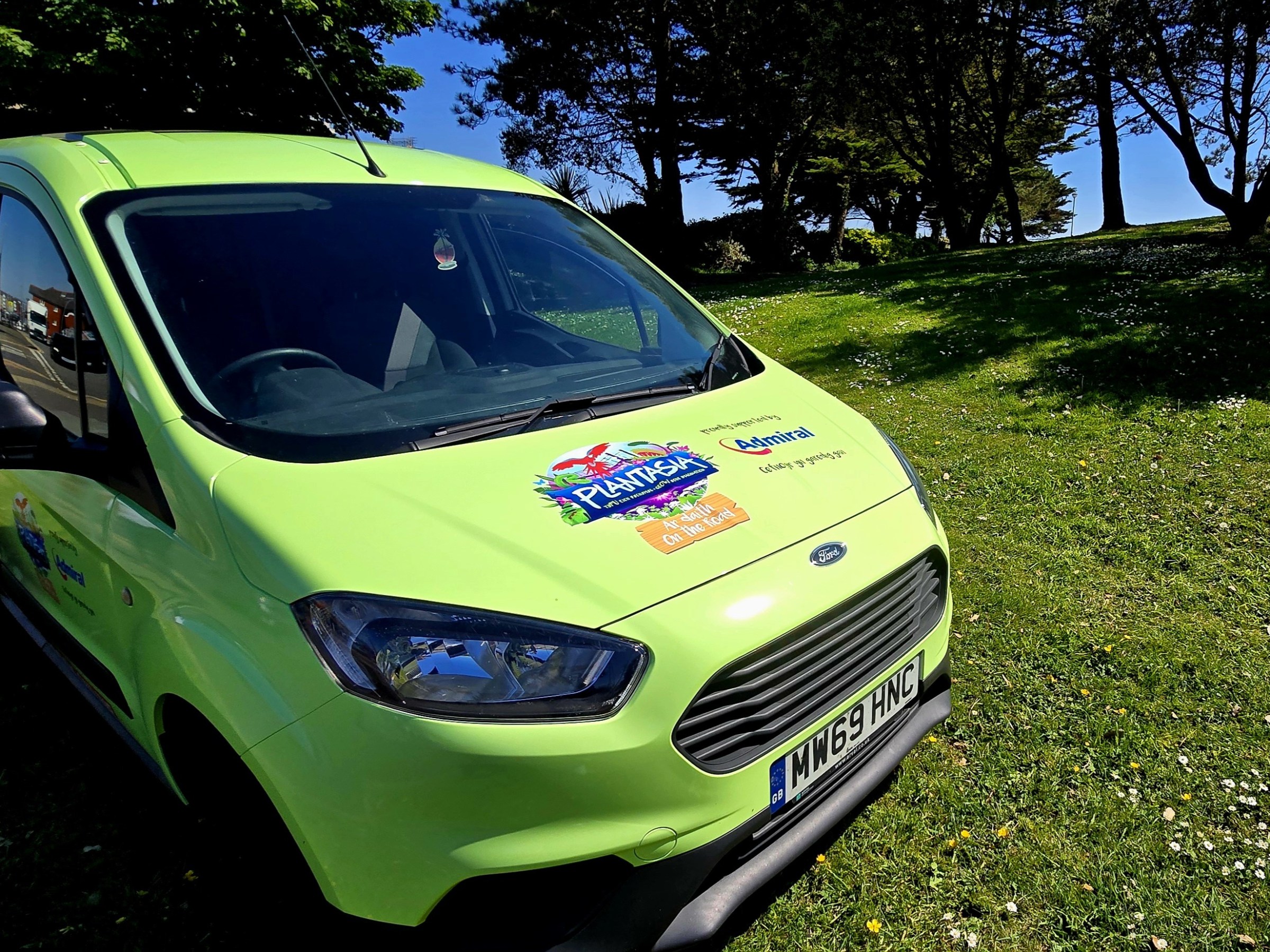 Lime green van with Plantasia on the Road logo parked on grass under trees on a sunny day.