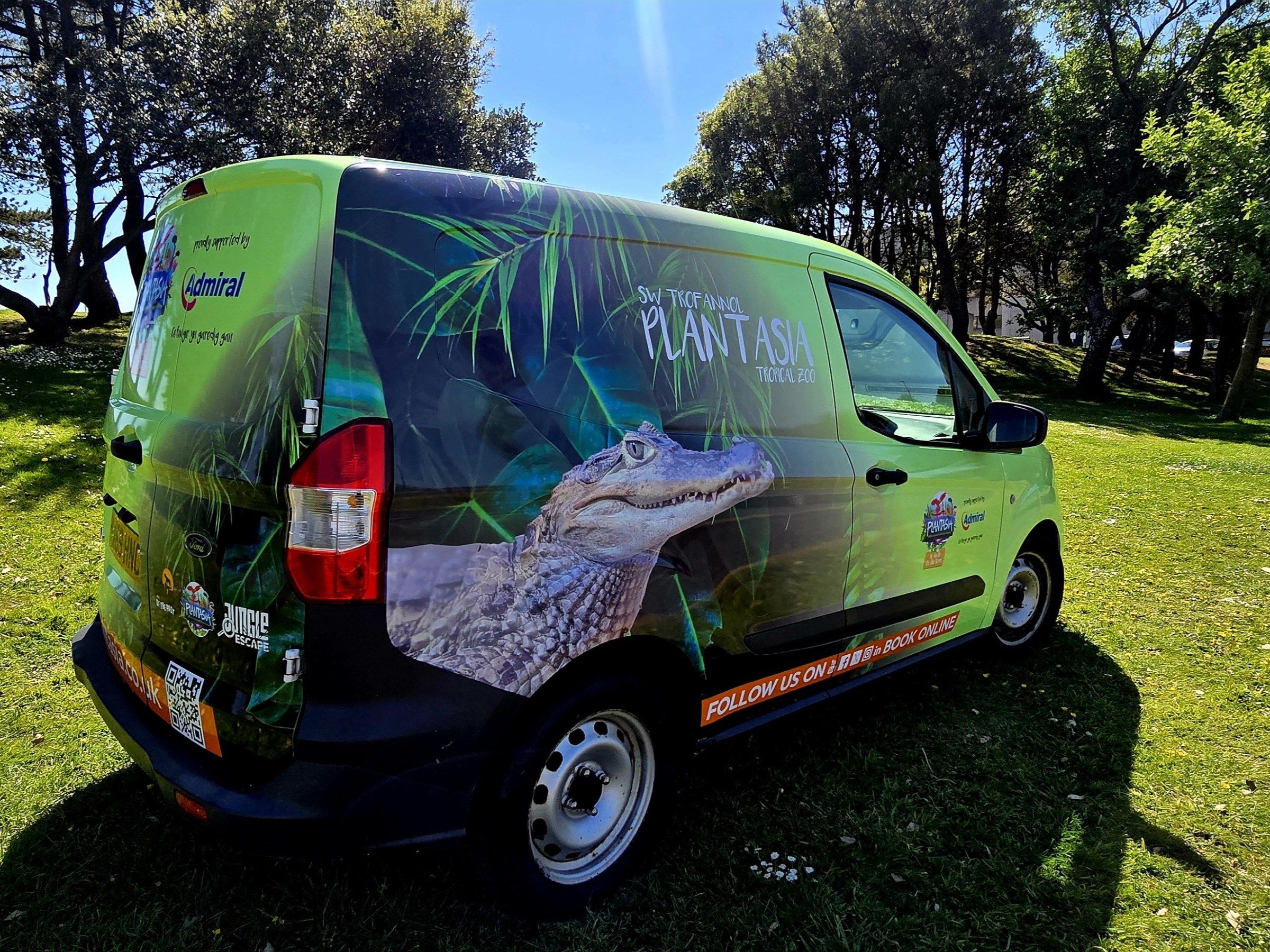 Green van with tropical zoo ads parked on grass near trees.