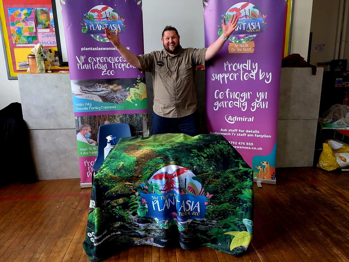 A male zookeeper standing with arms outstretched behind a table with Plantasia banners.