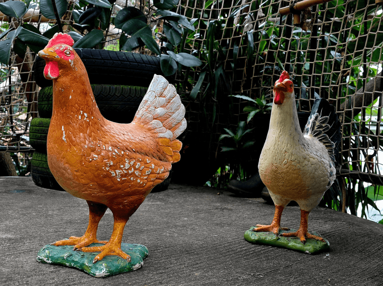 Two rooster statues on a concrete surface with foliage background.
