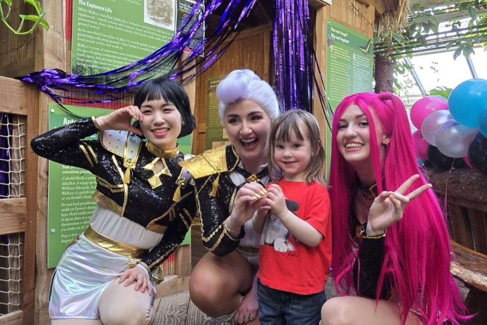 Three cosplayers with a child, posing happily in front of colorful backgrounds and balloons.