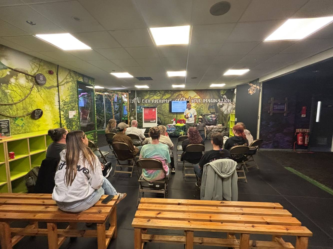 Group seated indoors, watching a presenter in a bright, green-themed room.